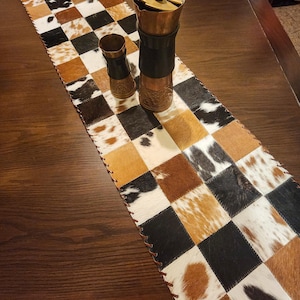 Peut inclure: Chemin de table en peau de vache avec un motif en damier dans les tons de marron, noir et blanc. Deux moulins à épices couleur cuivre sont posés sur le chemin de table. Le chemin de table est bordé d'une bordure en cuir cousue.