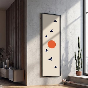 Könnte beinhalten: Ein gerahmter, vertikaler Kunstdruck mit minimalistischem Design. Das Kunstwerk zeigt einen cremefarbenen Hintergrund, eine orangefarbene Sonne und marineblaue Vögel im Flug. Der Rahmen ist schwarz.