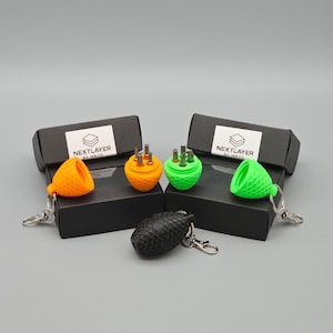 Könnte beinhalten: Eine Sammlung von 3D-gedruckten Schlüsselanhängern in Orange, Grün und Schwarz. Die Schlüsselanhänger sind an kleinen, schwarzen Schachteln mit dem Text "NEXTLAYER BY MR3D" auf der Vorderseite befestigt. Die Schlüsselanhänger haben ein einzigartiges, strukturiertes Design.