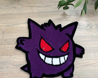 Puede incluir: Alfombra morada y negra con una imagen de dibujos animados de un Pokémon parecido a un fantasma con ojos rojos y una amplia sonrisa.