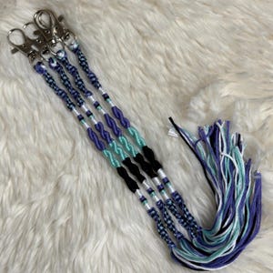 Peut inclure: Quatre porte-clés faits à la main avec des fermoirs argentés. Chaque porte-clés présente un motif tressé avec des sections alternées de fils bleus, turquoise, noirs et blancs. Un paquet séparé de fils assortis est également visible.