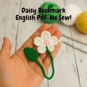 Op de afbeelding: Een handgemaakte gehaakte madeliefjesbladwijzer met een witte bloem, roze centrum en groene stengel en blad. De tekst "Daisy Bookmark English Pdf-No Sew!" wordt boven de bladwijzer weergegeven. De bladwijzer wordt in een hand gehouden.
