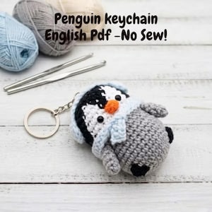 May include: Crocheted penguin keychain with a light blue scarf and orange beak. The penguin is attached to a silver keyring. The text "Penguin keychain English Pdf - No Sew!" is visible in the background.
