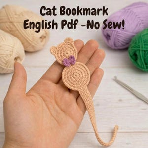 May include: A crocheted cat bookmark in beige with a small purple bow. The bookmark is held in a hand, with yarn balls in the background. Text on the image reads "Cat Bookmark English Pdf - No Sew!"