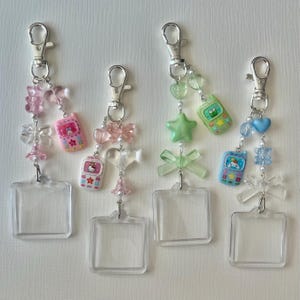 May include: Four keychains with clear plastic squares, silver clasps, and decorative charms. Charms include pink and blue cell phones, bows, stars, hearts, and beads. The keychains are arranged in a row against a white background.