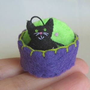 May include: A black felt cat with green eyes sits inside a purple felt cup with a green felt lining.