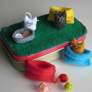 Puede incluir: Una escena caprichosa con figuras de gatos de fieltro y accesorios. Una base de fieltro verde se encuentra dentro de una lata con borde rojo, con un gato blanco, un gato negro y un gato naranja. Los accesorios incluyen una cama para gatos gris, una almohada amarilla y bolas de hilo.