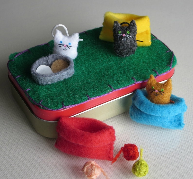 Puede incluir: Una escena caprichosa con figuras de gatos de fieltro y accesorios. Una base de fieltro verde se encuentra dentro de una lata con borde rojo, con un gato blanco, un gato negro y un gato naranja. Los accesorios incluyen una cama para gatos gris, una almohada amarilla y bolas de hilo.