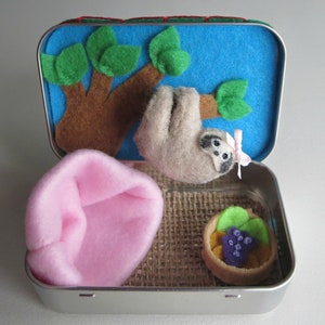 May include: A small tin box with a felt sloth hanging from a felt tree. The box also contains a pink felt cushion, a hessian fabric, and a felt dessert with purple berries.