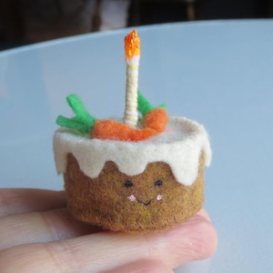 May include: A miniature felt birthday cake with white frosting, orange carrots, and a lit candle. The cake has a smiling face and is held in a hand. The candle has a yellow and orange flame.