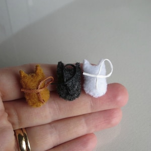 Puede incluir: Tres adornos de fieltro en miniatura con forma de gato en tonos naranja, negro y blanco. Cada gato tiene un dise&ntilde;o sencillo y cosido y un peque&ntilde;o lazo para colgar. El gato naranja tiene una cinta marr&oacute;n, el gato negro tiene una cuerda negra y el gato blanco tiene una cuerda blanca.