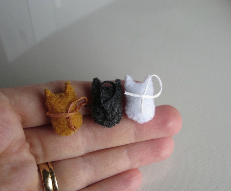 Puede incluir: Tres adornos de fieltro en miniatura con forma de gato en tonos naranja, negro y blanco. Cada gato tiene un dise&ntilde;o sencillo y cosido y un peque&ntilde;o lazo para colgar. El gato naranja tiene una cinta marr&oacute;n, el gato negro tiene una cuerda negra y el gato blanco tiene una cuerda blanca.