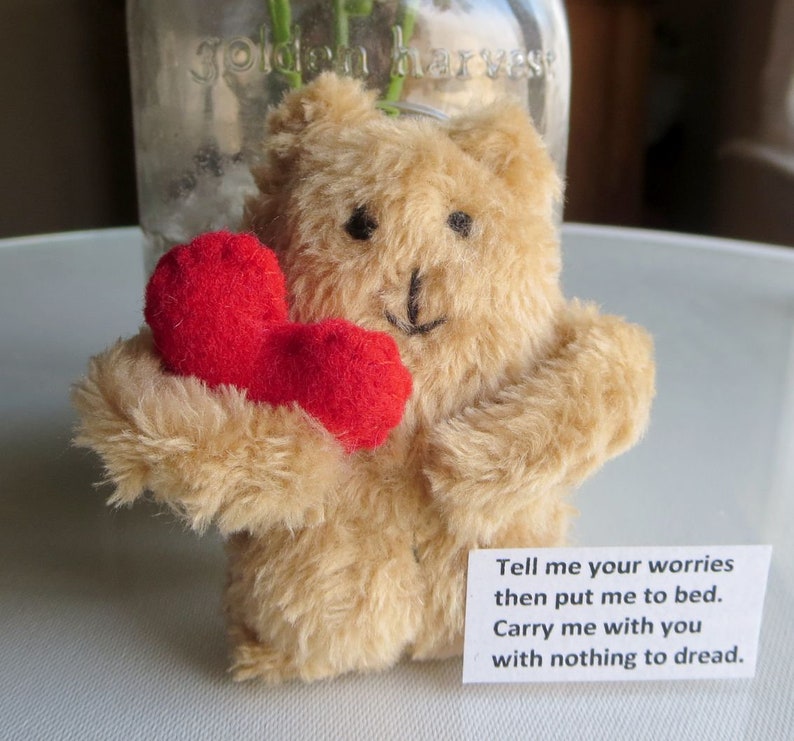 May include: A small, tan teddy bear holding a red heart. The bear has black button eyes and a stitched black mouth. A small white tag with the text "Tell me your worries then put me to bed. Carry me with you with nothing to dread" is attached.