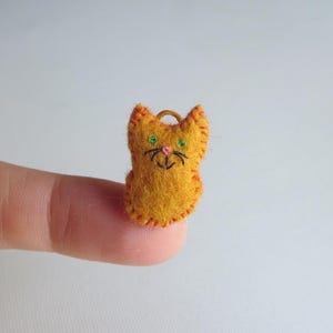 May include: A small, handmade orange felt cat with green eyes, a pink nose, and a black embroidered mouth and whiskers. The cat has orange stitching around the edges and a small loop at the top. The cat is a miniature craft item.