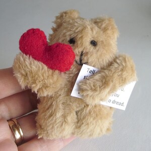 May include: A small, tan teddy bear holding a red heart and a small white card with text. The bear has black eyes and a black stitched nose. The heart is a vibrant red color. The bear's fur is soft and fluffy.