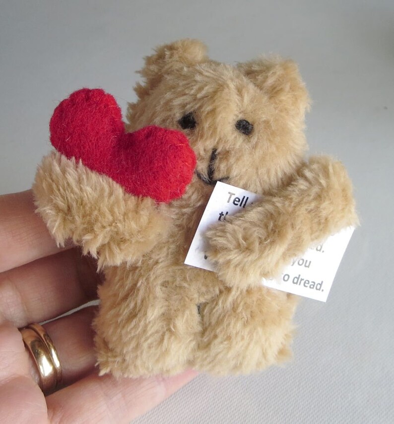 May include: A small, tan teddy bear holding a red heart and a small white card with text. The bear has black eyes and a black stitched nose. The heart is a vibrant red color. The bear's fur is soft and fluffy.