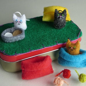 Puede incluir: Una escena de gato de fieltro caprichosa con una base de lata roja y una parte superior de fieltro verde. La escena incluye un gato blanco, un gato gris, un gato naranja en una manta azul, una bolsa roja y bolas de hilo. Un cuenco de comida de fieltro y una almohada amarilla completan la escena.