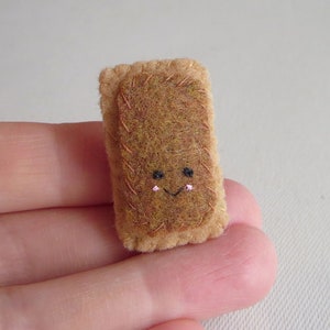 Puede incluir: Un alimento de fieltro marrón con una cara sonriente bordada. La cara tiene ojos negros y un rubor rosado.