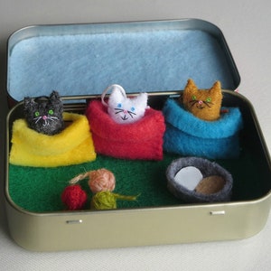 Puede incluir: Una caja de metal con interior azul alberga tres figuras de gato de fieltro en mantas amarillas, rojas y azules. Un peque&ntilde;o nido de fieltro con dos huevos y bolas de hilo tambi&eacute;n est&aacute; dentro. La caja est&aacute; sobre una superficie blanca.