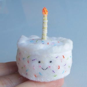 May include: A small, white felt cake with colorful sprinkles and a smiling face. A single candle with a yellow and orange flame sits atop the cake. The cake is a handmade item.
