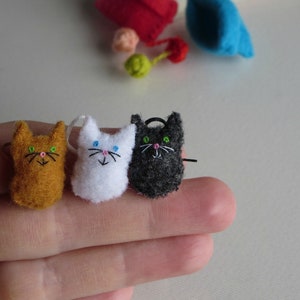 Puede incluir: Tres peque&ntilde;os adornos de fieltro con forma de gato en naranja, blanco y negro. Cada gato tiene rasgos faciales bordados, incluyendo bigotes y una nariz rosa. Los gatos se muestran en una mano, con otras formas de fieltro coloridas en el fondo.