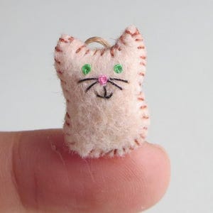May include: A small, handmade felt cat ornament. The cat is light pink with brown stitched details, green eyes, a pink nose, and black whiskers. A loop of twine is attached for hanging.