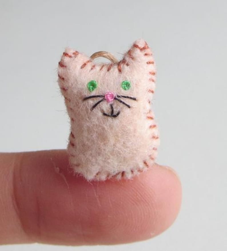May include: A small, handmade felt cat ornament. The cat is light pink with brown stitched details, green eyes, a pink nose, and black whiskers. A loop of twine is attached for hanging.