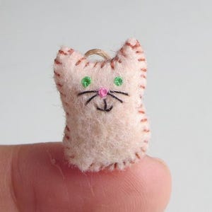 May include: A small, handmade felt cat ornament. The cat is light pink with brown stitching around the edges and on the ears. It has green eyes, a pink nose, and black whiskers. A loop of twine is attached for hanging.
