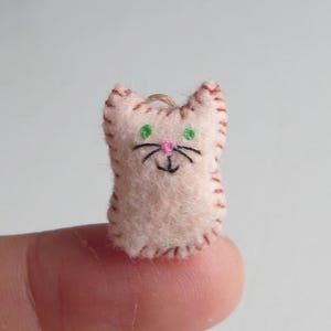 May include: A small, handmade felt cat ornament. The cat is light pink with brown stitched edges, green embroidered eyes, a pink nose, and black whiskers. A loop of twine is attached for hanging.