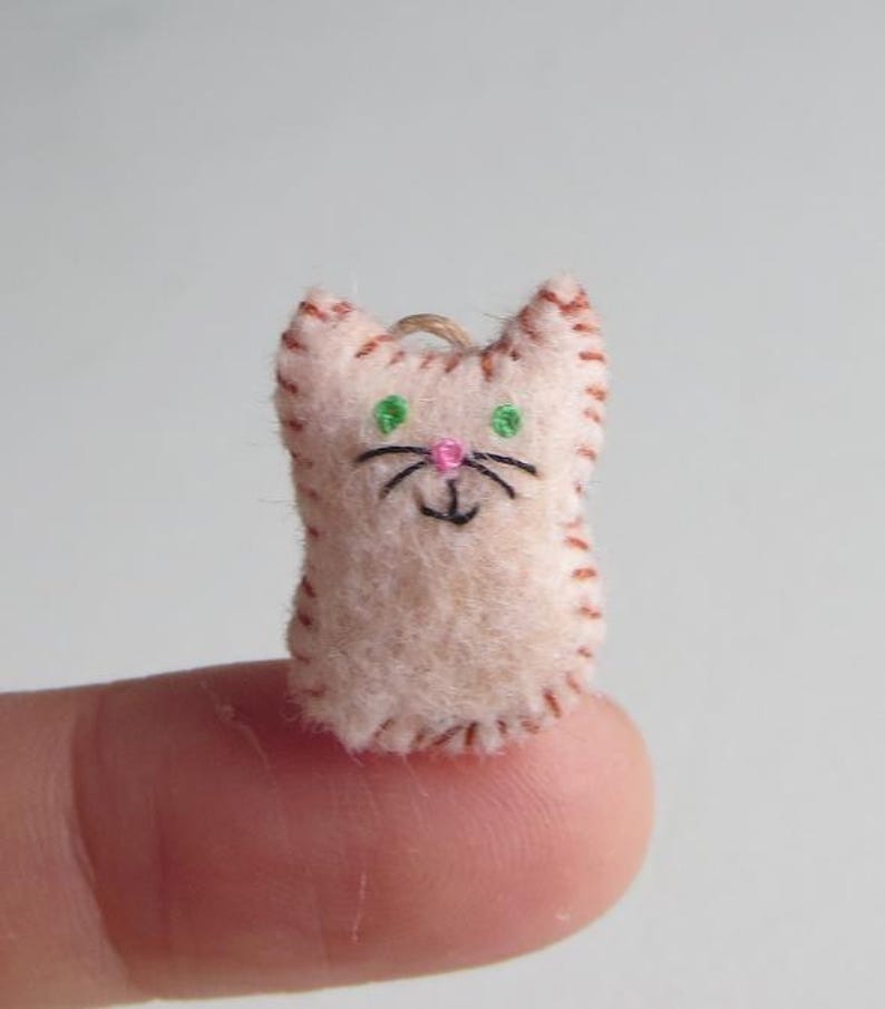May include: A small, handmade felt cat ornament. The cat is light pink with brown stitched edges, green embroidered eyes, a pink nose, and black whiskers. A loop of twine is attached for hanging.