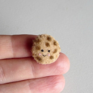 May include: A small, brown, felt cookie with a smiling face and chocolate chips.