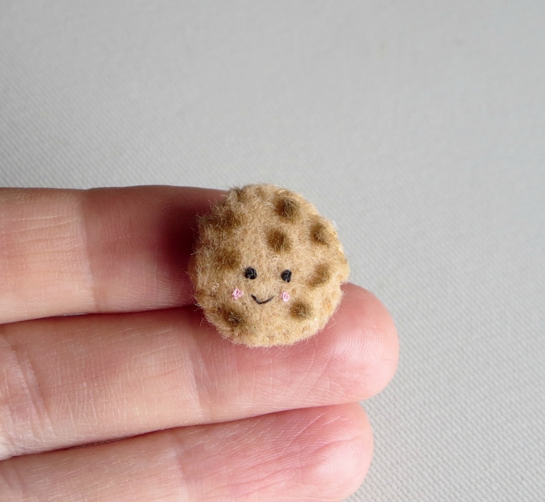 May include: A small, brown, felt cookie with a smiling face and chocolate chips.