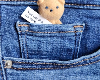 May include: A small, tan, needle-felted bear figurine peeking out of a blue denim pocket. Next to the bear is a white paper note with the printed text, "You got this. I'm here." The bear has two dark blue stitched eyes and a small nose.