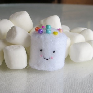 May include: A white felt marshmallow with a smiling face and colorful sprinkles on top. The marshmallow is surrounded by white marshmallows.