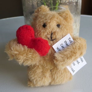 May include: A small, tan teddy bear holding a red felt heart. The bear has black button eyes and a stitched smile. Two small white tags are attached to the bear with handwritten text. The bear is a handmade item.