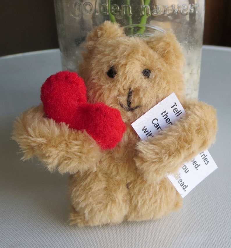 May include: A small, tan teddy bear holding a red felt heart. The bear has black button eyes and a stitched smile. Two small white tags are attached to the bear with handwritten text. The bear is a handmade item.