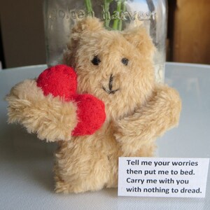 May include: A tan teddy bear holding two red hearts. The bear has black eyes and a stitched black nose and mouth. A small white card with the text "Tell me your worries then put me to bed. Carry me with you with nothing to dread." is attached.