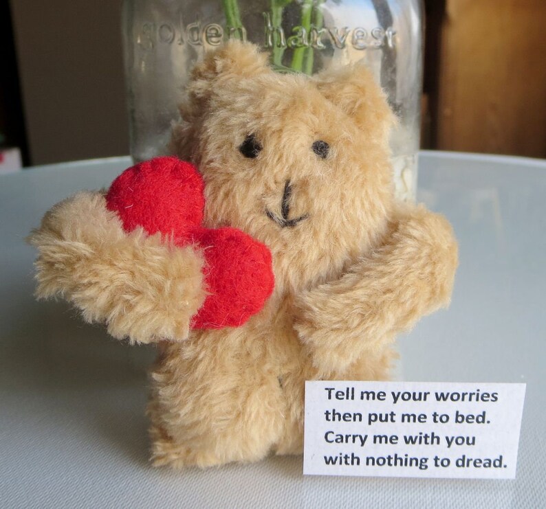 May include: A tan teddy bear holding two red hearts. The bear has black eyes and a stitched black nose and mouth. A small white card with the text "Tell me your worries then put me to bed. Carry me with you with nothing to dread." is attached.