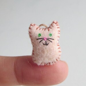 May include: A small, handmade felt cat figurine. The cat is light pink with brown stitched details, green eyes, and a pink nose. It has a loop at the top for hanging. The cat is approximately 1 inch tall.