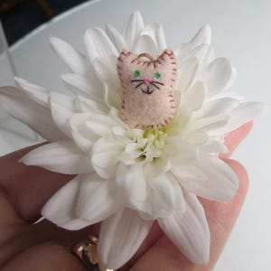 May include: A small, light pink felt cat-shaped ornament with embroidered details, including green eyes and a black nose and whiskers. The cat is perched on a large, white chrysanthemum flower, creating a whimsical contrast.