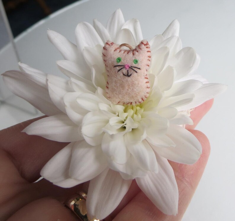 May include: A small, light pink felt cat-shaped ornament with embroidered details, including green eyes and a black nose and whiskers. The cat is perched on a large, white chrysanthemum flower, creating a whimsical contrast.
