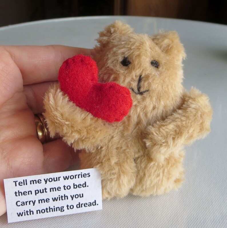 May include: A small, tan teddy bear holding a red heart. The bear has black eyes and a stitched smile. A small white card with the text "Tell me your worries then put me to bed. Carry me with you with nothing to dread." is visible.