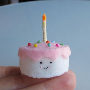 Könnte beinhalten: Ein kleiner, handgefertigter Geburtstagskuchen aus Filz mit einer rosa Zuckerguss-Schicht und bunten Streuseln. Obenauf befindet sich eine gelb-orangefarbene Kerze. Der Kuchen hat ein lächelndes Gesicht.