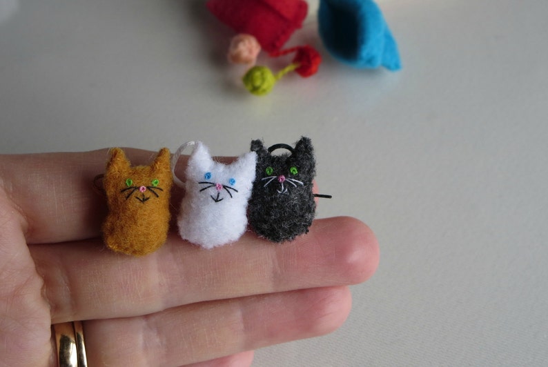 Puede incluir: Tres peque&ntilde;os adornos de gato de fieltro hechos a mano en naranja, blanco y negro. Cada gato tiene rasgos faciales bordados, incluyendo ojos, nariz y bigotes. El gato naranja tiene ojos verdes, el gato blanco tiene ojos azules y el gato negro tiene ojos verdes.