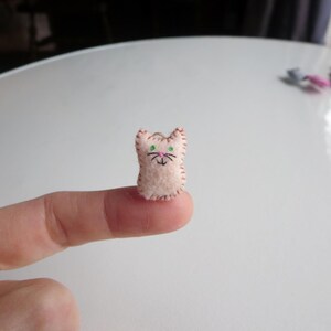 May include: A small, handmade felt cat figurine. The cat is light pink with embroidered details, including green eyes, a pink nose, and black whiskers. The edges are stitched with a contrasting thread. The cat is approximately 0.75 inches tall.