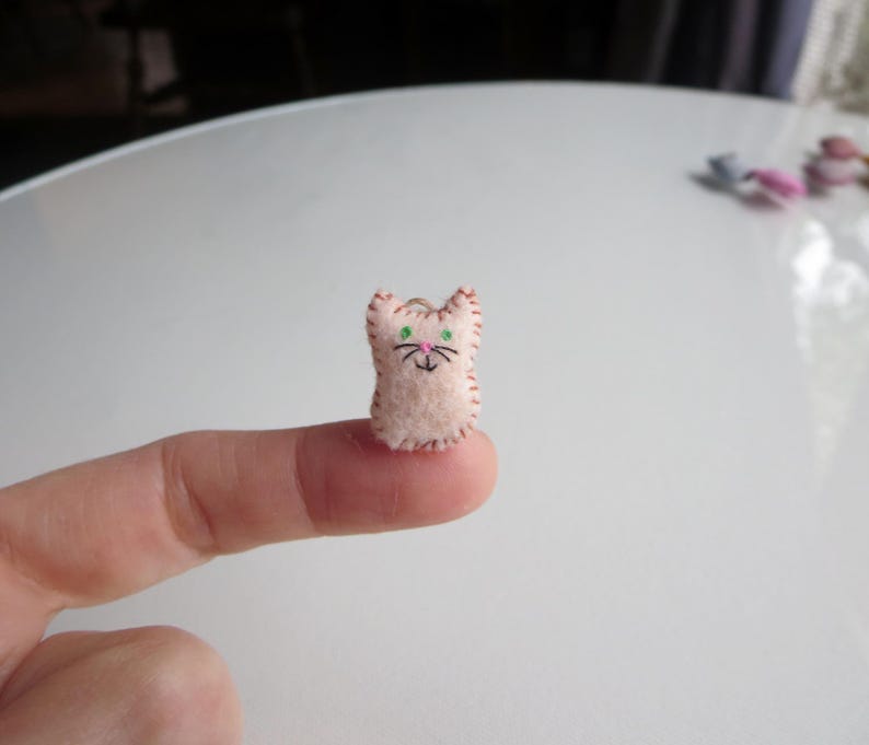 May include: A small, handmade felt cat figurine. The cat is light pink with embroidered details, including green eyes, a pink nose, and black whiskers. The edges are stitched with a contrasting thread. The cat is approximately 0.75 inches tall.