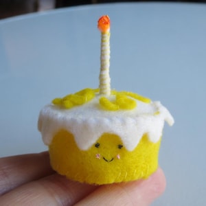 May include: A small, handmade felt birthday cake with a yellow base, white frosting, and a single candle. The cake has a smiling face and yellow decorative accents. The candle has an orange flame.