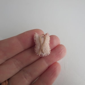 May include: A small, handmade felt ornament in the shape of a heart. The ornament is light pink with a brown stitched outline and a brown twine design. The heart is held in a hand.