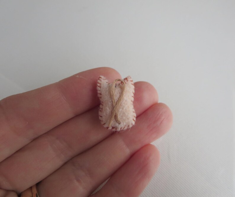 May include: A small, handmade felt ornament in the shape of a heart. The ornament is light pink with a brown stitched outline and a brown twine design. The heart is held in a hand.
