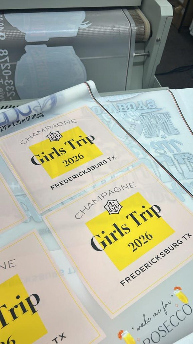 May include: A sheet of printed heat transfer decals featuring rectangular labels with the text CHAMPAGNE, Girls Trip, 2026, and FREDERICKSBURG TX in black font over a yellow background, alongside other graphic designs on a white backing material.